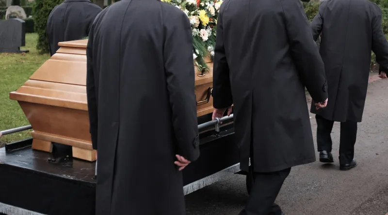 Mehrere Sargträger begleiten einen Holzsarg auf einem Friedhof zur Beisetzung.